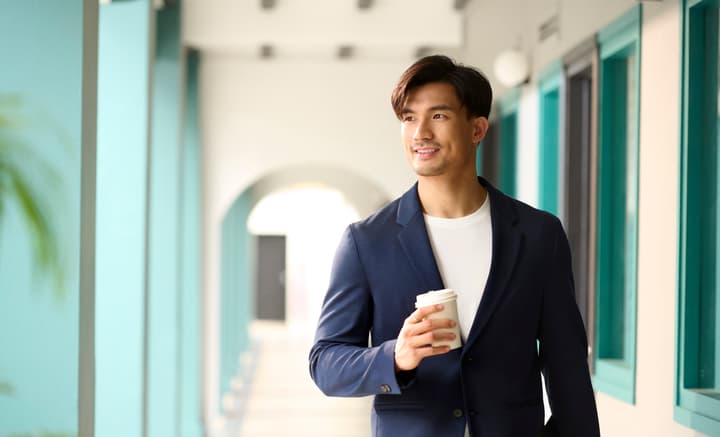 Male resident in a blazer walking down the covered corridor of the historic shophouse, carrying a coffee cup, with teal columns framing the walkway.