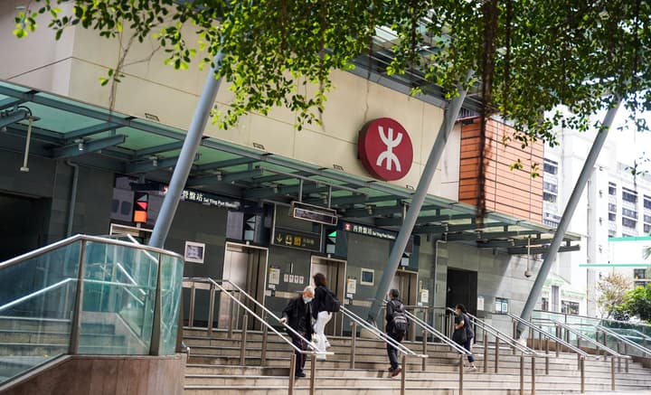 Exterior of the Sai Ying Pun MTR station entrance in Hong Kong, showing the red MTR logo and people ascending the outdoor stairs.