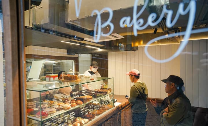 A glass storefront window of a bakery, displaying pastries and showing customers placing an order at the counter.
