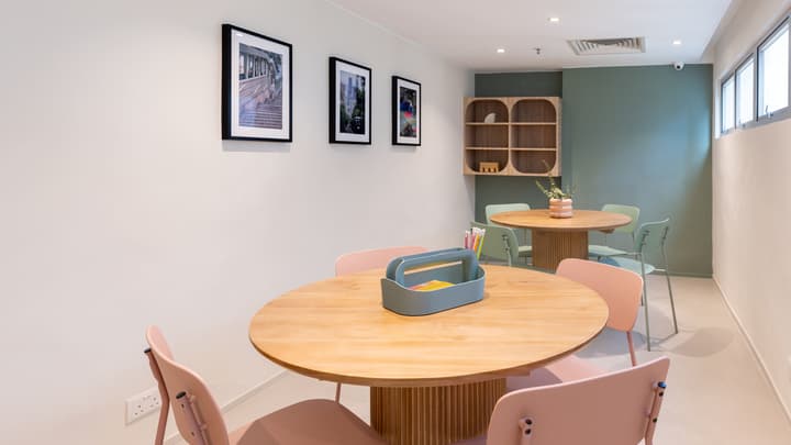 Bright communal workspace featuring two wooden round tables with pastel pink and mint-green chairs, set against a white and olive-green wall with framed photos.