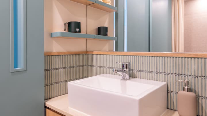 Modern bathroom close-up showing a square sink, vertical pastel blue tile, a mirrored wall, and open shelves holding dark green and black cups.