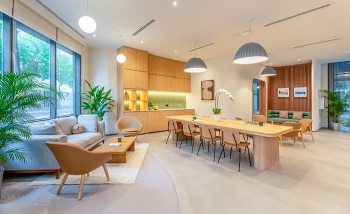 Bright communal lounge and co-working area featuring a long wooden communal table, various seating areas, and a small kitchenette with light wood cabinets.