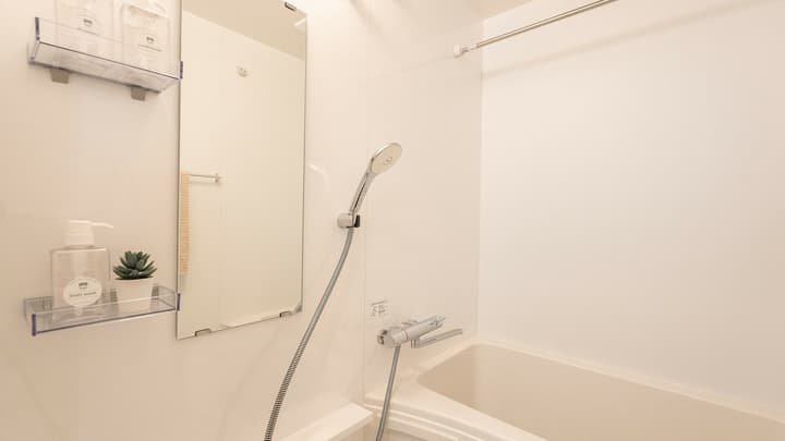 Compact, all-white Japanese unit bathroom featuring a deep bathtub, a hand-held shower head, a large wall mirror, and clear acrylic storage shelves.
