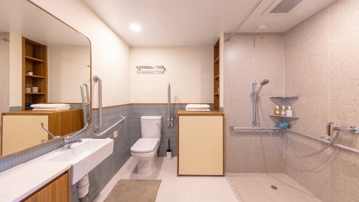 Spacious and accessible bathroom featuring a wall-mounted sink, grab bars next to the toilet and in the roll-in shower area, and light blue and speckled beige tile.