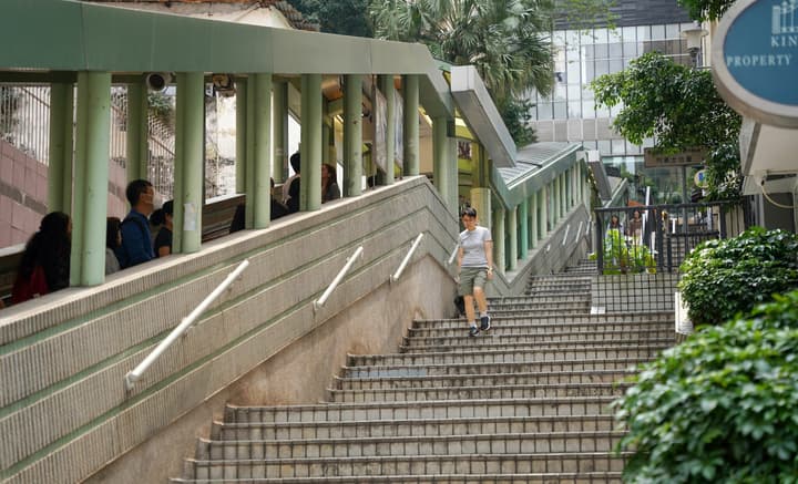 A person walking down a long, stepped public pathway with a green covered escalator structure running alongside it in an urban setting.