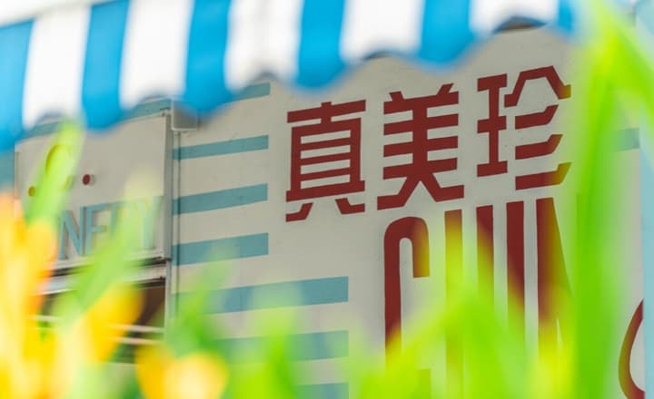 Close-up, blurred shot through green foliage of the red Chinese characters for Chin Mee Chin Confectionery painted on a white wall under a blue-and-white striped awning.