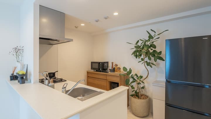 Compact, modern kitchen with a black refrigerator, white countertops, sink, single cooktop, microwave on a wooden cabinet, and a large potted plant.