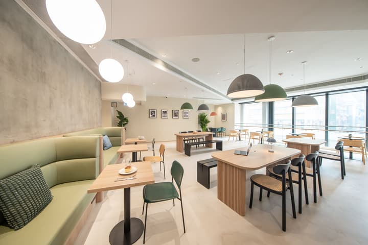 A wide view of a bright, modern communal lounge or cafe area with large windows. The seating is a mix of long light-green upholstered booths, small tables for two, and long communal wooden tables with black and cane chairs.