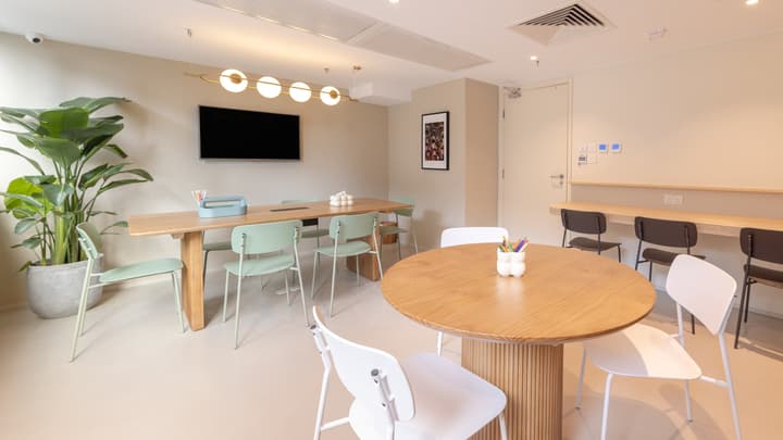 Multi-purpose room with a long wooden table for meetings or group work, a smaller round table, mint-green and white chairs, and a wall-mounted monitor.