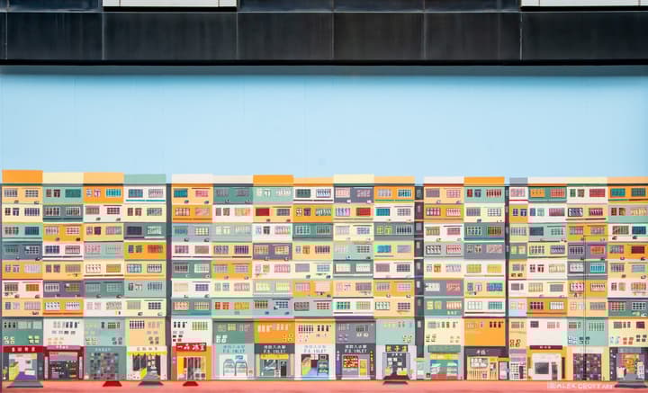 A wide, flat mural depicting a dense block of colorful apartment buildings and street-level shops in a highly geometric, repeating pattern against a plain blue sky background.