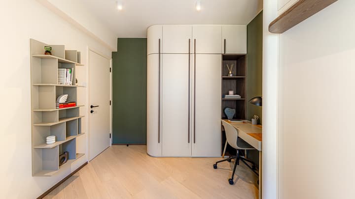 Flexible apartment room set up as a study with a sleek desk, curved built-in wardrobe, floor-to-ceiling shelving, and a dark green accent wall.