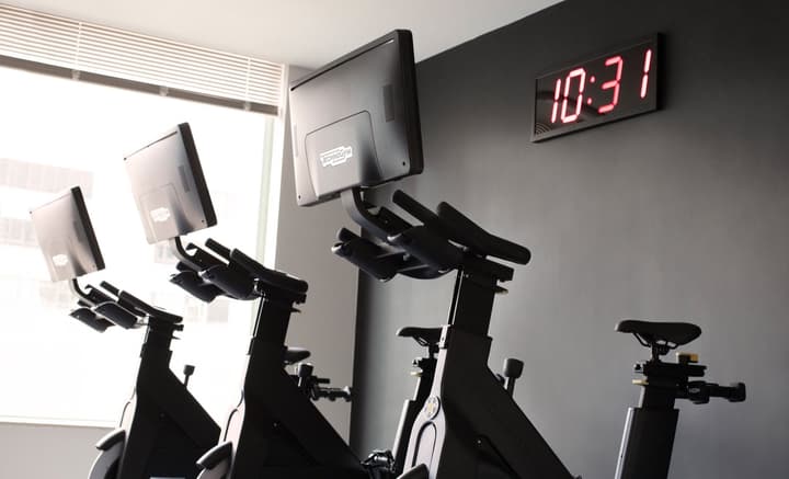 A close-up view of black spin bicycles with mounted screens in a modern fitness studio, set against a dark gray wall with a large digital clock displaying "10:31."