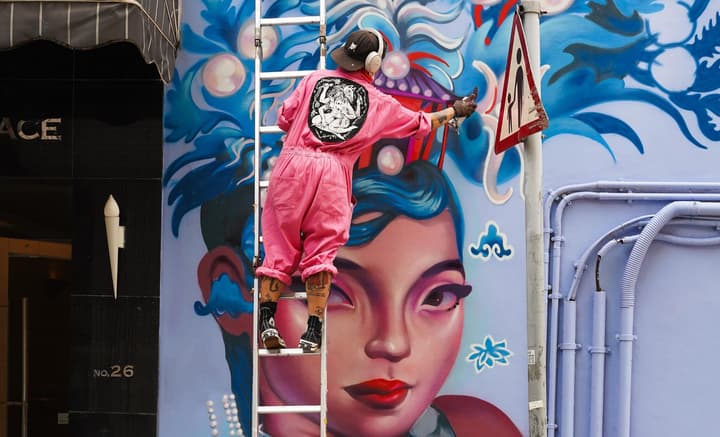 A street artist in a pink jumpsuit and headphones painting a large, vibrant blue and pink mural of a woman's face on a city wall.