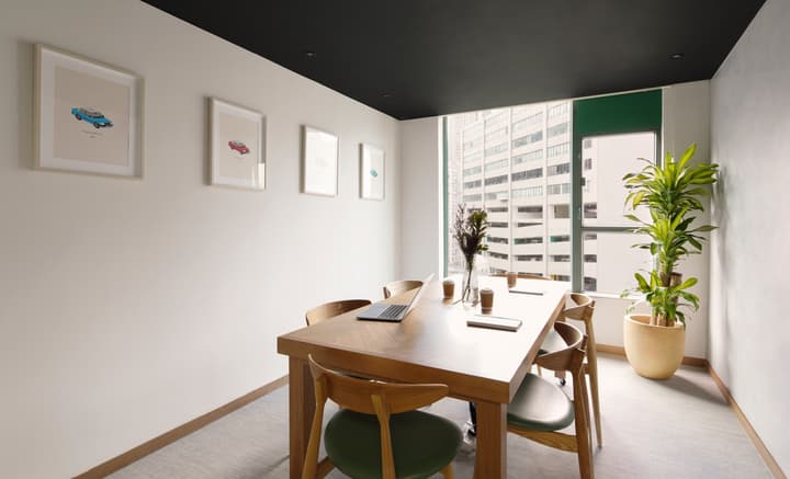 A brightly lit private meeting room with a wooden table, green-seated chairs, white walls with simple framed art, and a tall window offering a view of the adjacent buildings.