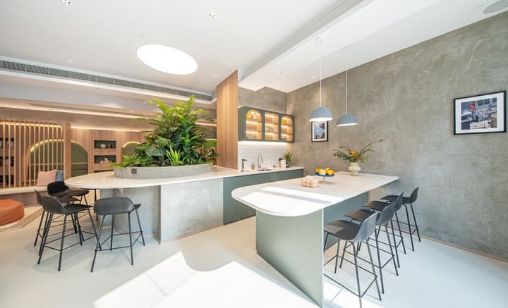View of a modern kitchen area centered around a large oval island with black bar stools. One section of the island features a built-in planter with lush green foliage, while another section acts as a counter next to green cabinets against a textured gray wall.