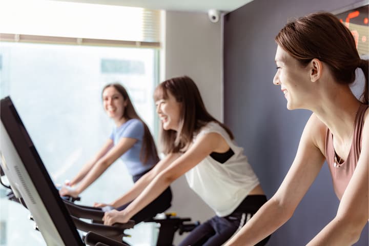 Three happy women smiling while exercising together on spin bikes in a brightly lit indoor fitness or gym area.