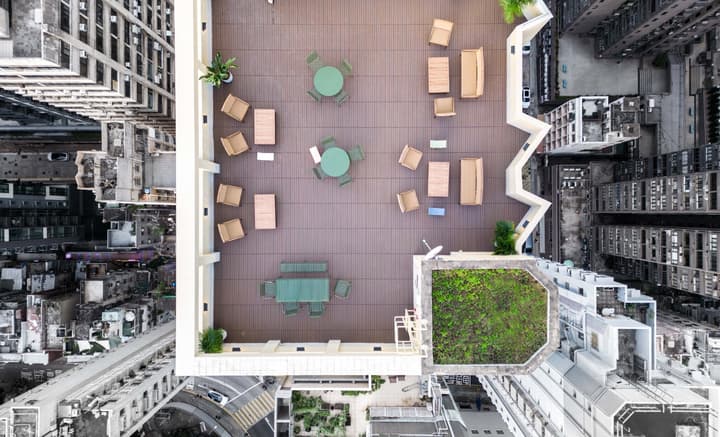 Aerial view of a rectangular, wood-decked rooftop terrace with various outdoor seating arrangements and green patches, surrounded by tall city buildings.