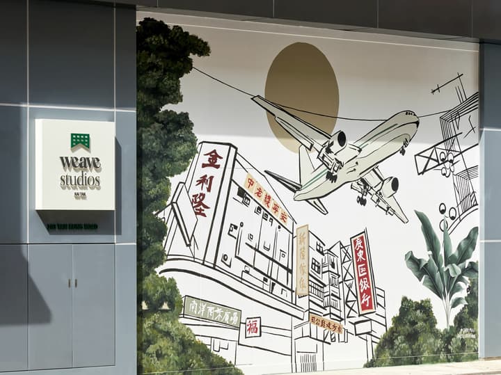 Exterior view of the "Weave Studios Kai Tak" sign next to a large mural depicting an airplane landing over Hong Kong street buildings.