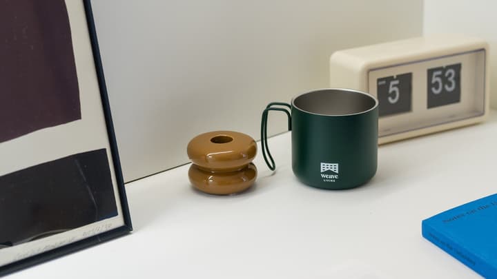Close-up of a desk surface with a green metal mug, a brown hourglass-shaped ceramic object, a vintage flip clock, and framed artwork.