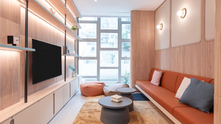 Sleek communal living room with wood wall paneling, an orange leather sofa, a built-in entertainment unit with light-up shelves, and windows overlooking the city.
