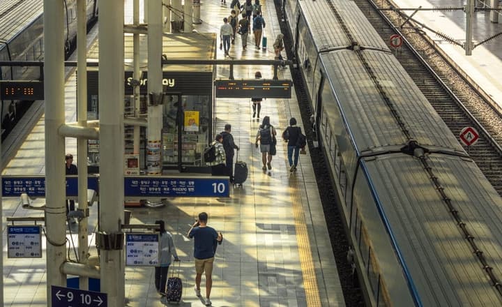 Overhead view of a busy Seoul train station platform (possibly subway or KTX) with commuters walking, a train stopped at the platform, and station signs visible.