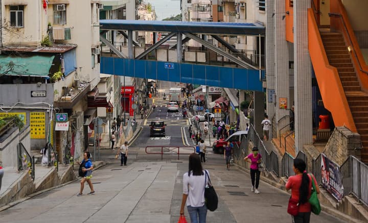 A view down a steep, busy Hong Kong street with pedestrians and cars, featuring a raised blue metal footbridge crossing overhead.