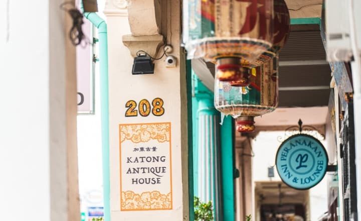 Close-up of the Katong Antique House sign on a white wall, with colorful Peranakan-style lanterns and a Peranakan Inn & Lounge sign hanging nearby.