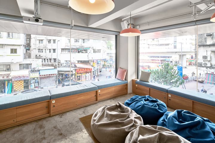 A cozy, bright communal lounge area with built-in wooden bench seating along a wide window, plush bean bag chairs in blue and beige, overlooking a busy city street.
