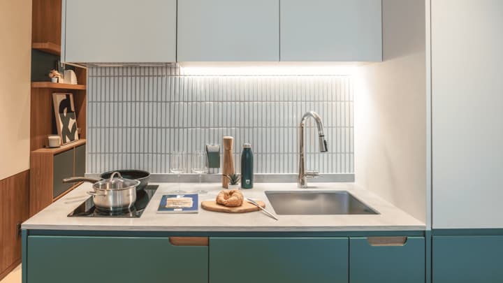 Stylish, modern kitchenette with deep green lower cabinets, light gray upper cabinets, a stainless steel sink, and a backsplash made of narrow, vertically oriented white tiles. A pan and pot are on the cooktop.