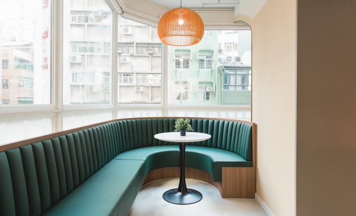 A cozy, curved window nook featuring built-in, deep teal velvet banquette seating with vertical channeling, a small white pedestal table, and an orange woven pendant light hanging overhead.