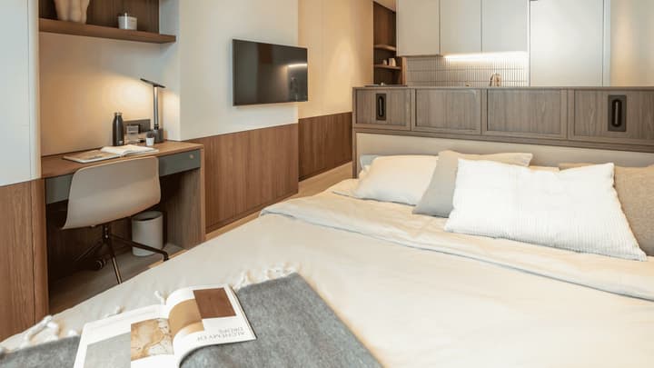 Interior view of the Queen Suite from the foot of the bed, showing the integrated desk and shelving area on the left and the compact kitchenette separated by a half-wall divider.