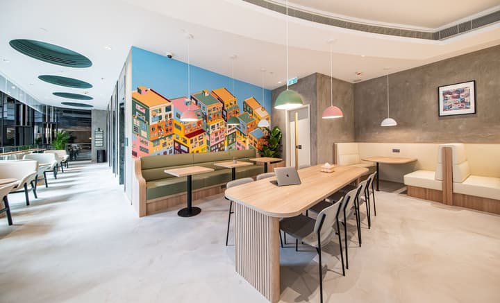 Bright, modern communal lounge area with a long wooden table for working or dining, flanked by built-in green and beige booth seating. A colorful wall mural of clustered houses brightens the space.