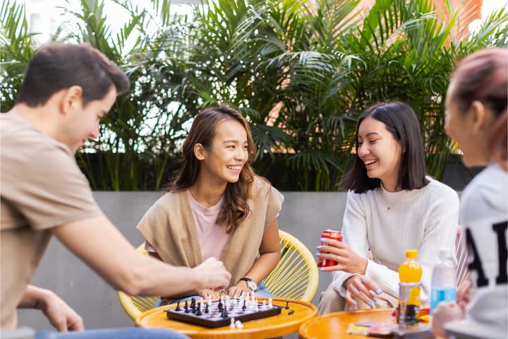 A group of four young friends sitting around a small table on a sunny outdoor terrace, smiling and playing a game of travel chess against a backdrop of dense green foliage.