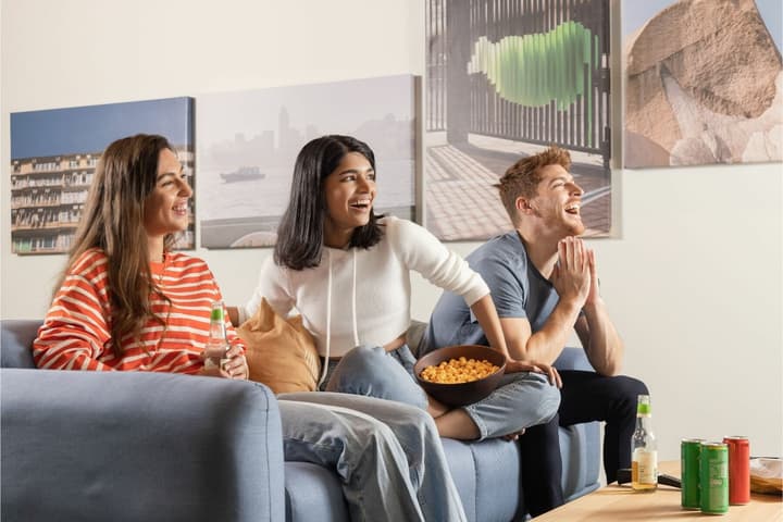 Three friends, two women and one man, laughing and enjoying snacks on a blue couch in a modern communal living room.
