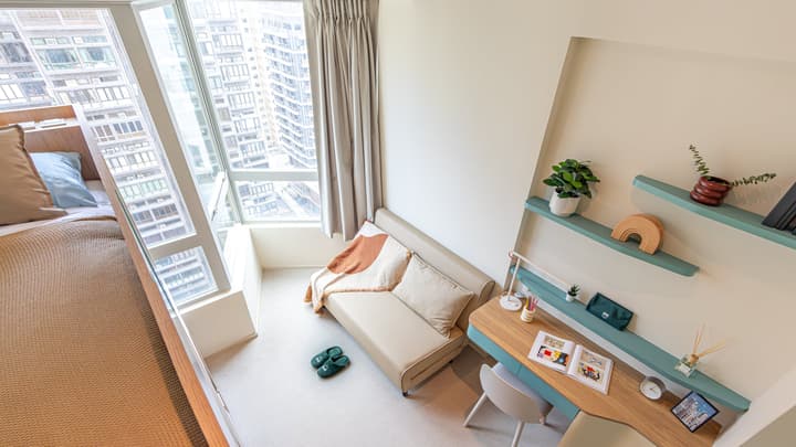 High-angle view of a studio apartment showing the bed, a small sofa, and a wooden desk area with mint-green shelving near a large angled window overlooking the city.