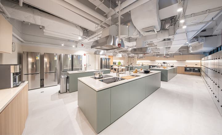 A spacious, commercial-style shared kitchen with multiple light green prep islands and stainless steel range hoods overhead. The room features several stainless steel refrigerators along one wall and a long wall of numbered storage lockers on the right. Exposed white ceiling ducts are visible above.