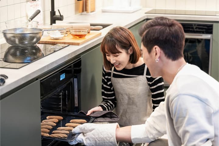 A smiling couple, a man and a woman, taking a tray of freshly baked cookies out of a modern oven in a shared coliving kitchen.