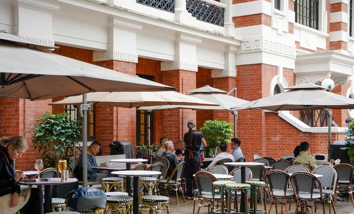 Busy outdoor café seating area with umbrellas and tables, set against a classic red brick and white trimmed historic building facade.