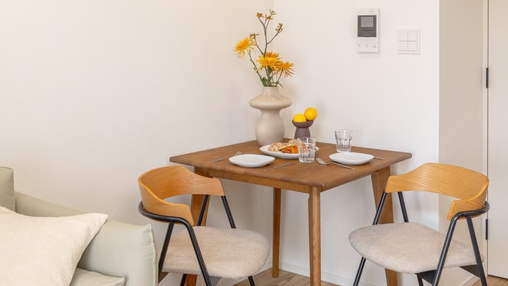 Close-up of a cozy, bright dining area with a two-person wooden table, minimalist mid-century style chairs, and a vase with yellow flowers.