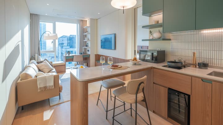 Wide view of the One Bedroom Luxe Suite showing the open kitchen with dark green cabinets and the living room separated by a half-wall, featuring a sofa lounge and a desk by the window.