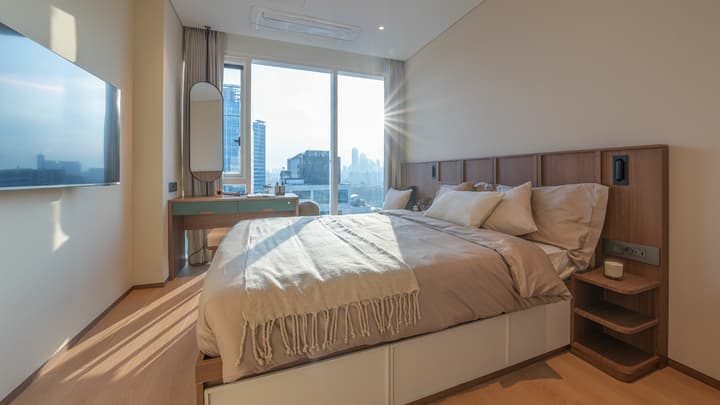 The separate bedroom of the luxe suite, featuring a queen bed with under-bed storage, a stylish wood headboard with integrated lighting, and a window-facing dressing table/desk.