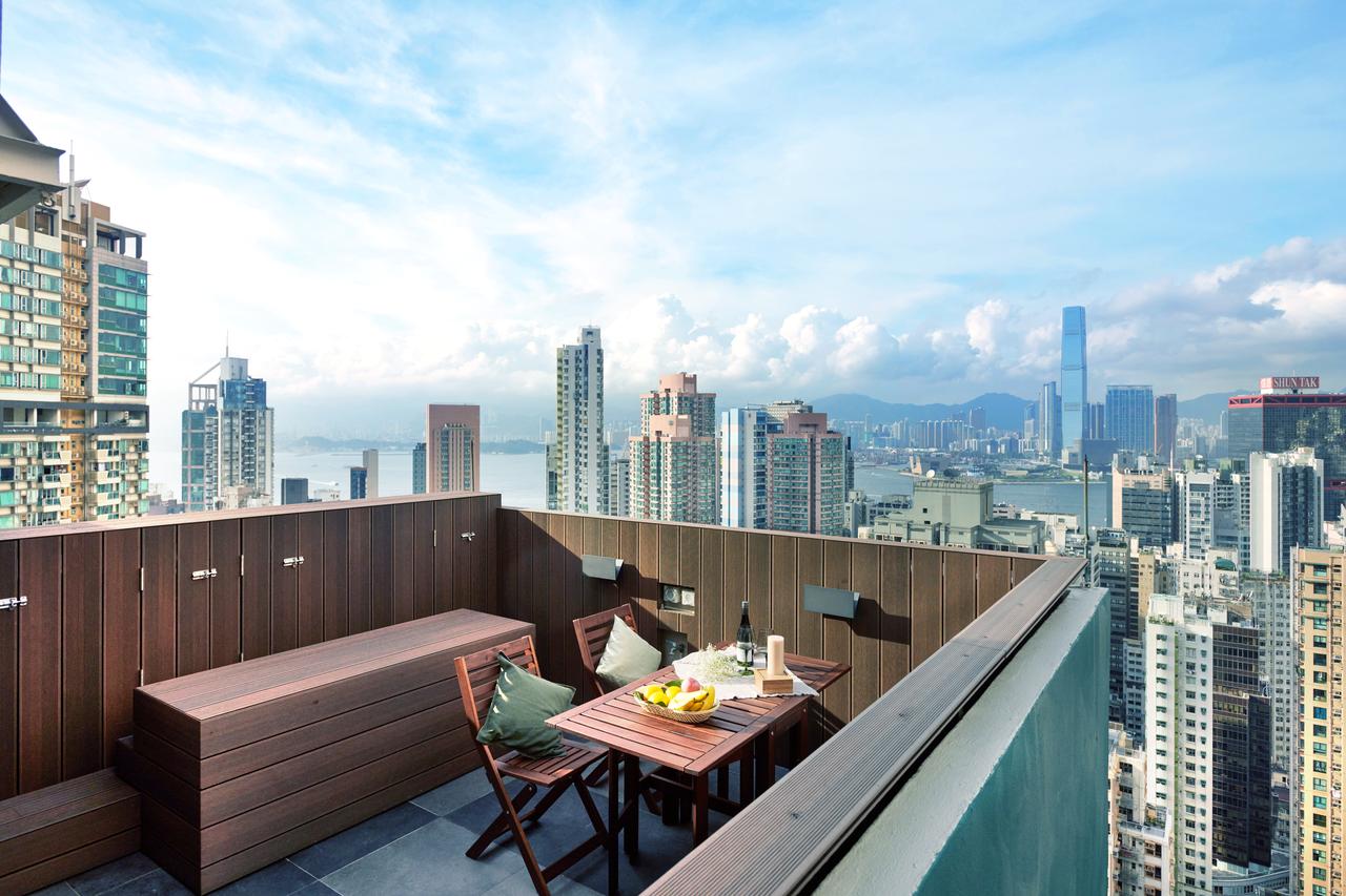 Stunning rooftop terrace with a wooden dining set and built-in bench, offering a panoramic view of the Hong Kong city skyline and harbor under a blue sky.