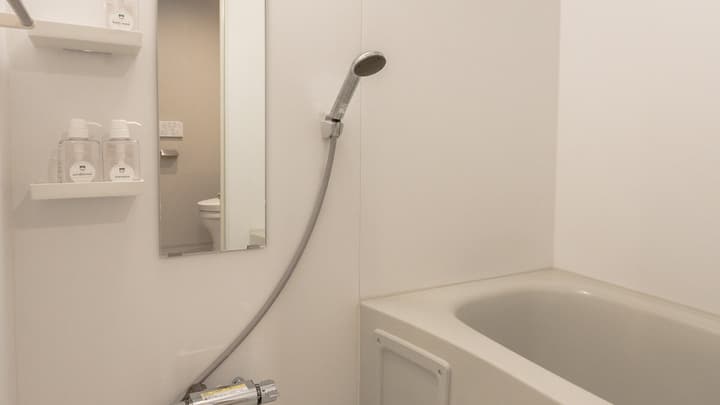 Interior view of the white, compact Japanese-style bathroom unit, showing the bathtub, a shower head, and wall-mounted shelving with body wash and shampoo bottles.