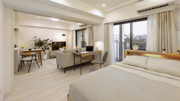 Wide view of a bright, open-concept studio apartment featuring a bed, work desk, couch, dining table, and balcony access.