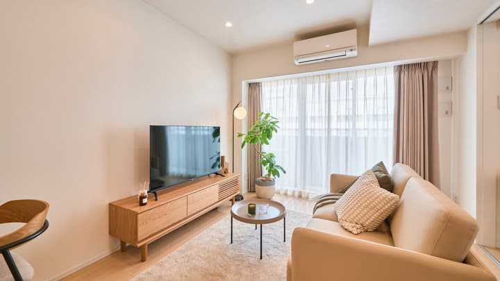 Minimalist living room with a cream-colored sofa, a wooden TV console, a large flat-screen TV, hardwood floors, and bright natural light from a full-wall window.