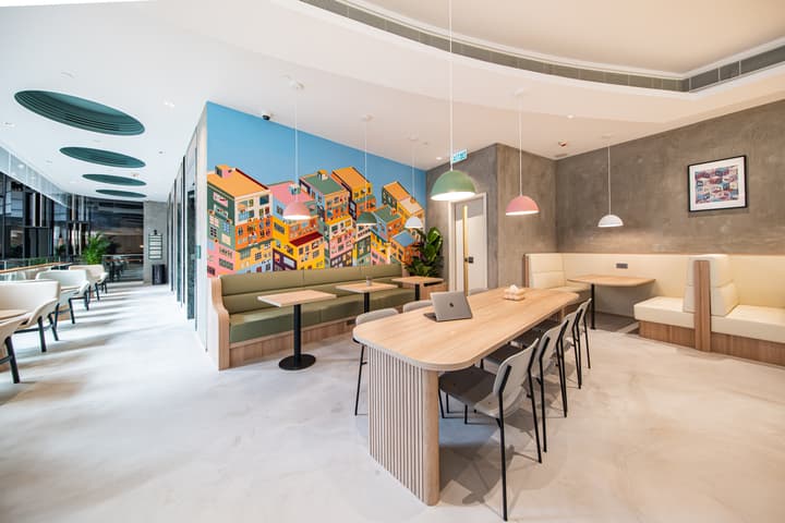 Bright, modern communal lounge area with a long wooden table for working or dining, flanked by built-in green and beige booth seating. A colorful wall mural of clustered houses brightens the space.