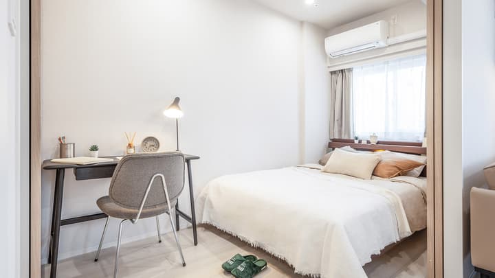Bedroom with a double bed, light-colored bedding, a bedside window, and a dedicated workspace featuring a dark desk, gray chair, and desk lamp.