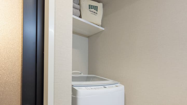 Close-up of the laundry area with an in-unit white washing machine and a shelf above it with folded towels and a storage bag.