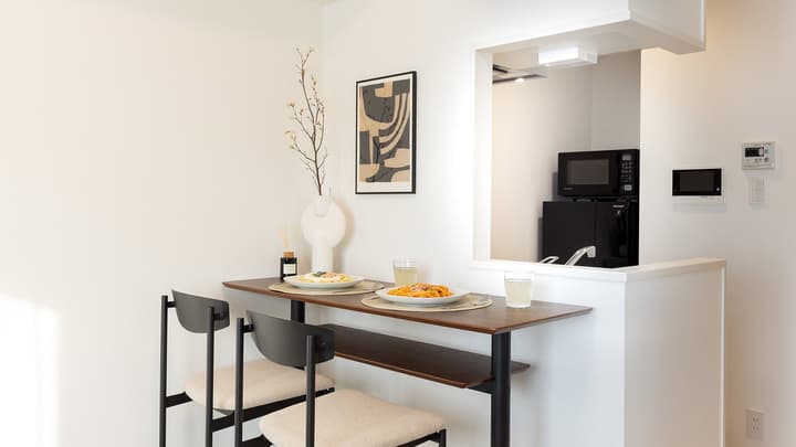 A two-seater dining bar table set for a meal, with a white vase and art, looking through a pass-through window into the kitchenette.
