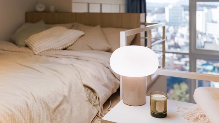 Close-up of a cozy loft sleeping area with a bed, soft linens, a modern glowing lamp on a nightstand, and a blurred city view from a window.